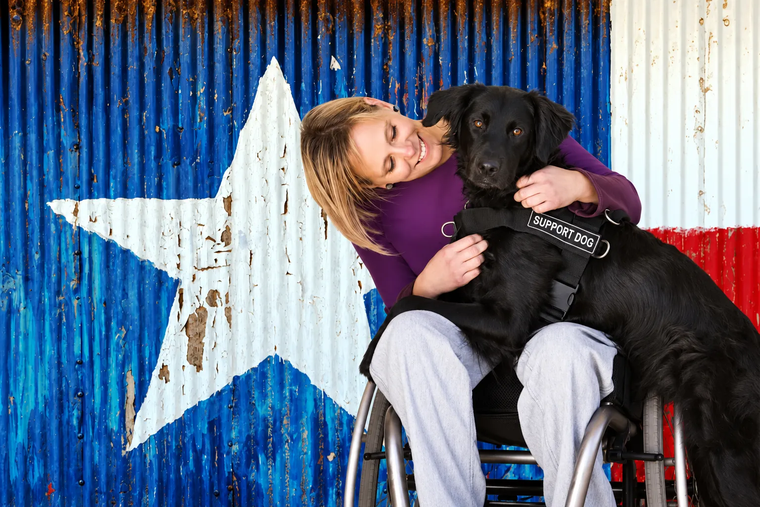 A woman using a wheelchair with her black support dog beside a Texas flag